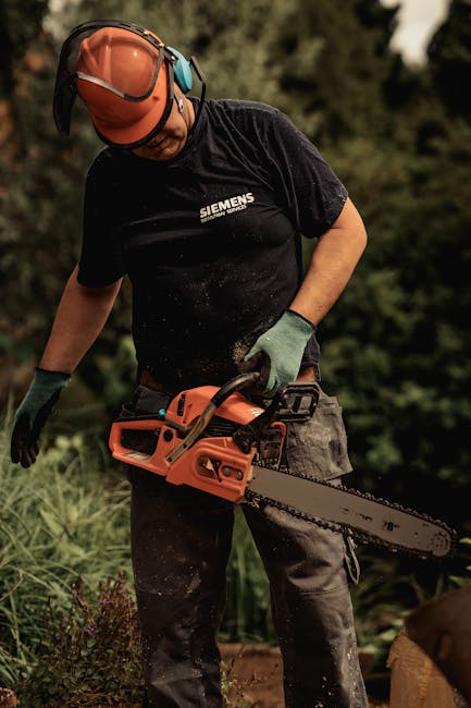 A man in a blue t-shirt is engaged in hedge trimming in a lush garden, holding a manual hedge trimmer with a long, serrated metal blade. The hedge is dense with vibrant green foliage, and the trimmed 